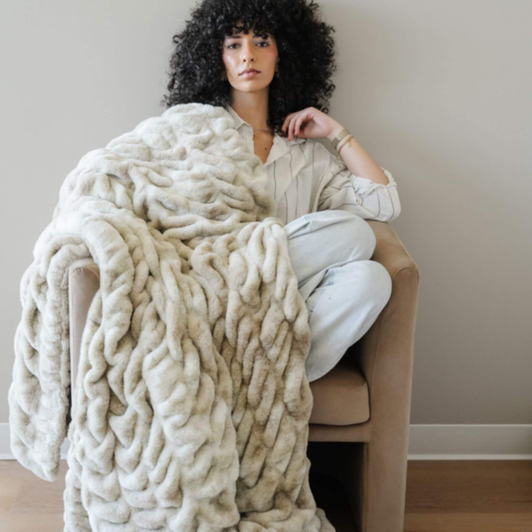 Woman sitting on a chair with Saranoni Double Ruched Faux Fur Throw Blankets Sand draped over her