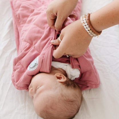 Baby in Dream Weighted Sleep Swaddle - Rose being adjusted by an adult on a white surface