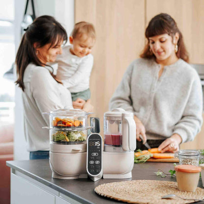 Two women and a child in a kitchen with Babymoov Duo Meal Station Baby Food Maker on the counter.