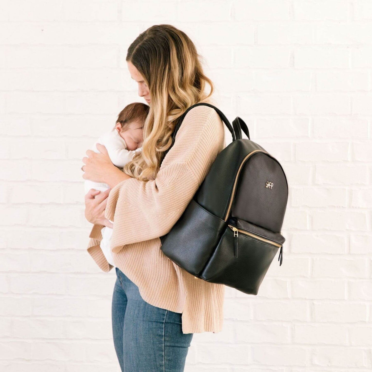 Woman holding a baby and carrying a Ebony Classic City Diaper Bag II against a white brick wall.
