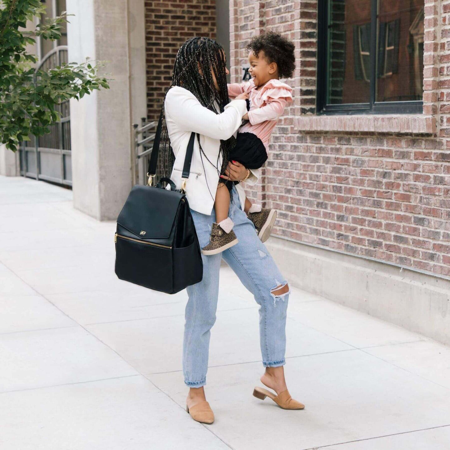 Woman holding a child and carrying Ebony Classic Diaper Bag II on a sidewalk.