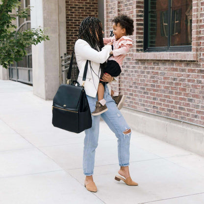 Woman holding a child and carrying Ebony Classic Diaper Bag II on a sidewalk.