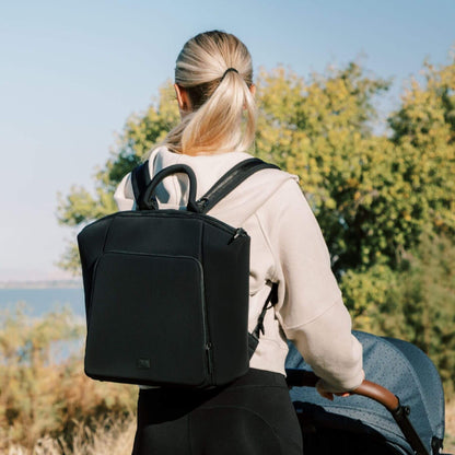 Person with Ebony Seoul Neoprene Backpack II walking outdoors with trees and clear sky in the background