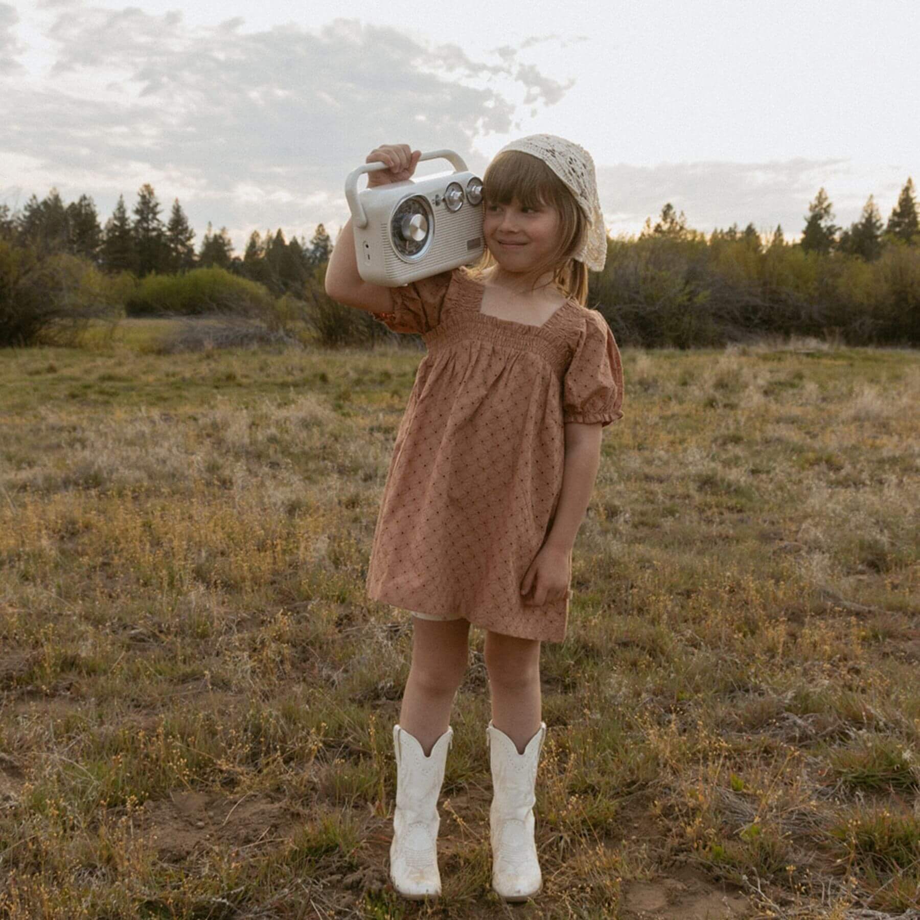 A young girl stands in an open grassy field holding a vintage-style white radio. She is wearing Makemake Organics Organic Eyelet Bubble Sleeve Dress - Clay Eyelet