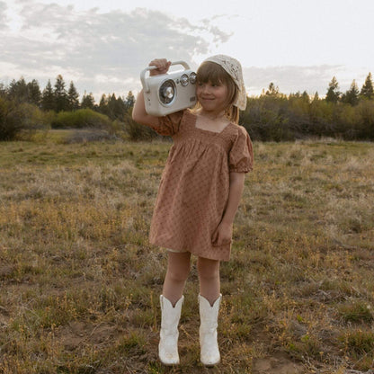 A young girl stands in an open grassy field holding a vintage-style white radio. She is wearing Makemake Organics Organic Eyelet Bubble Sleeve Dress - Clay Eyelet
