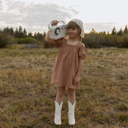 A young girl stands in an open grassy field holding a vintage-style white radio. She is wearing Makemake Organics Organic Eyelet Bubble Sleeve Dress - Clay Eyelet