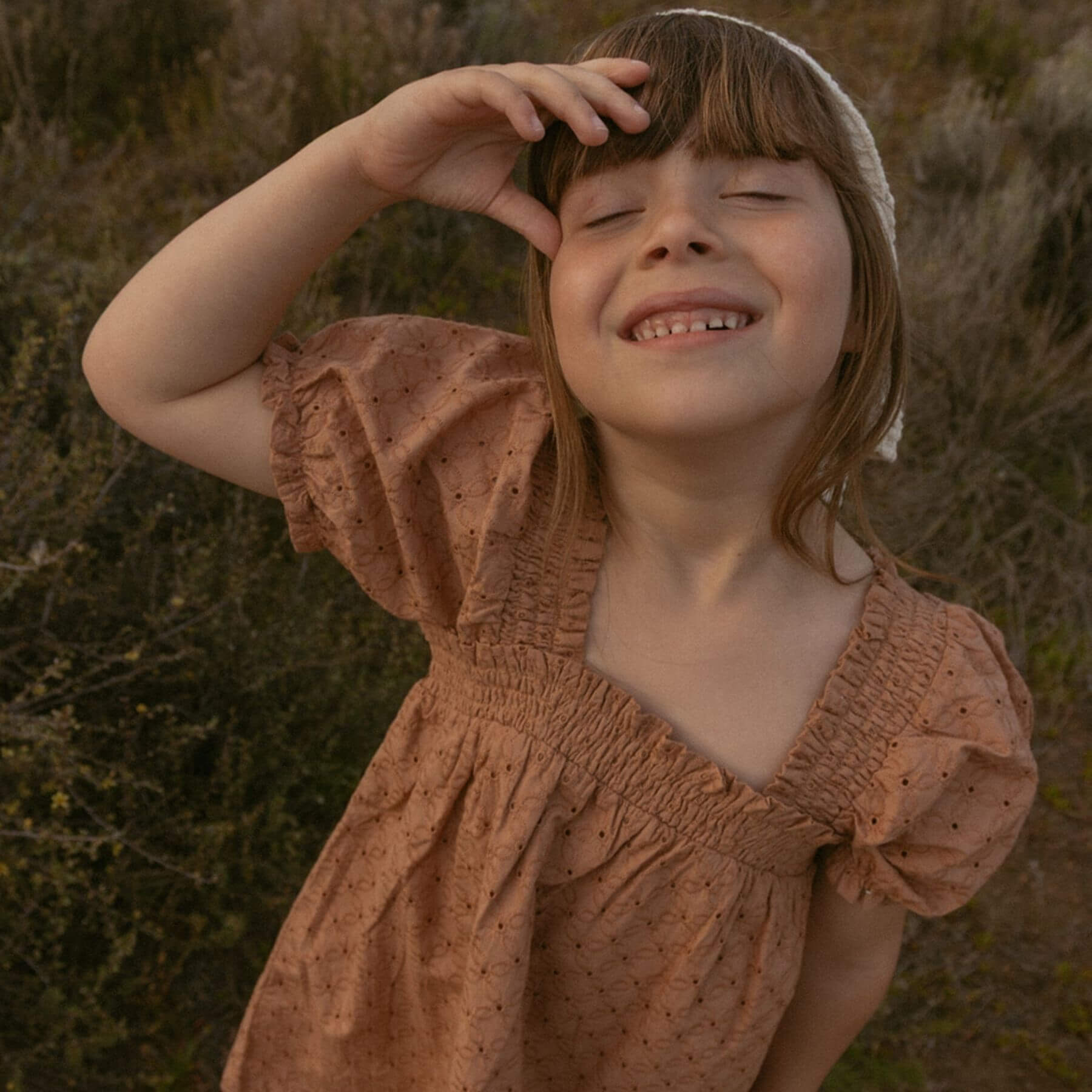 A young girl wearing Makemake Organics Organic Eyelet Bubble Sleeve Dress - Clay Eyelet with a white bonnet, smiling outdoors against a blurred natural background.