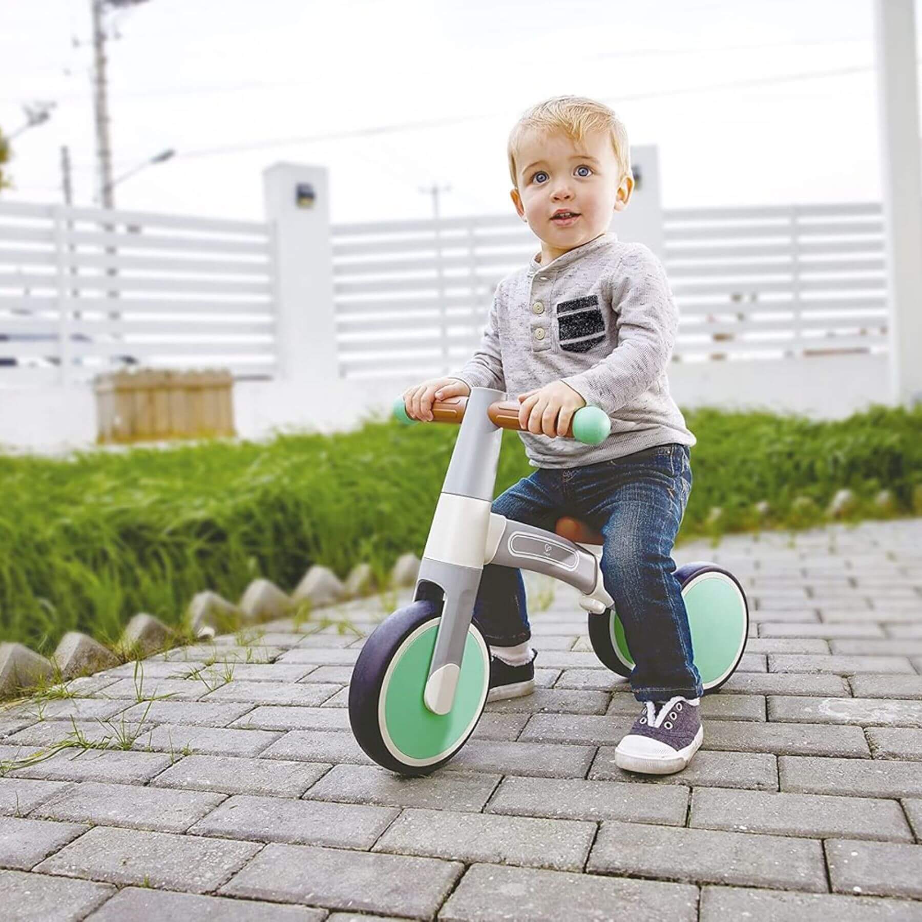 Child riding Hape First Ride Balance Bike Light Green on a paved path with grass and a white fence in the background