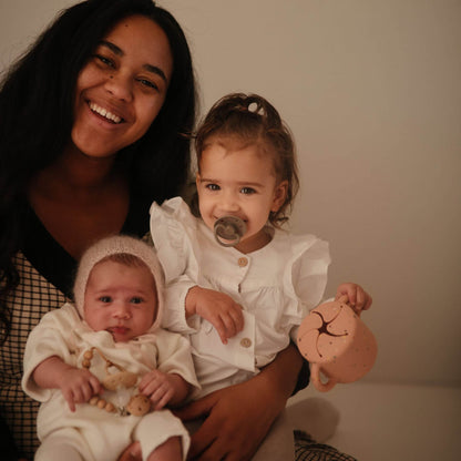 Woman holding two young children, one of whom is holding a toy, against a plain background.
