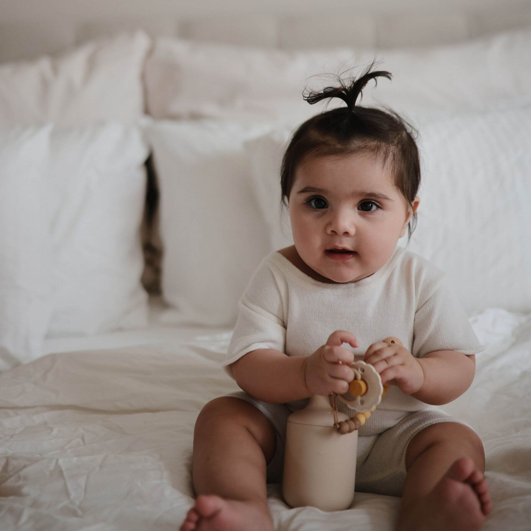 Baby sitting on a bed holding a FRIGG pacifier, wearing a white onesie.