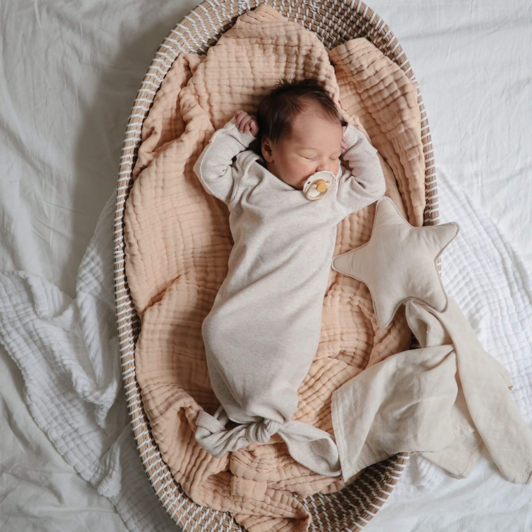 Newborn baby wrapped in a white blanket with a FRIGG pacifier, lying in a wicker basket with star-shaped pillow.