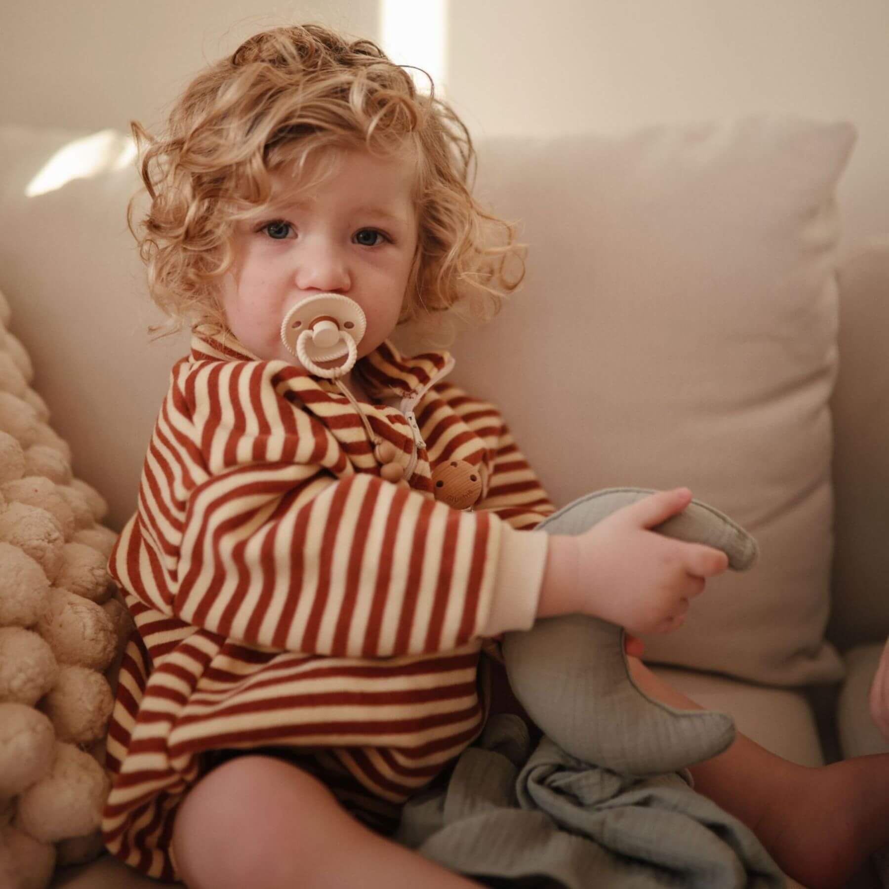 Child in a striped outfit holding a toy, sitting on a couch with a FRIGG pacifier in mouth.