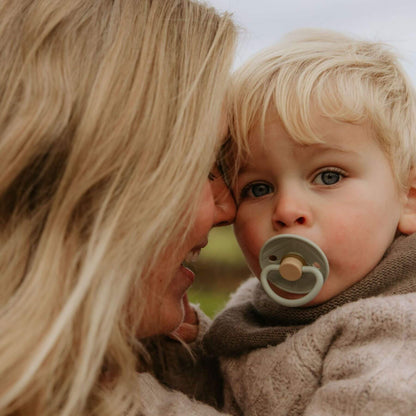 Woman holding a child with a FRIGG pacifier, both looking at each other outdoors.