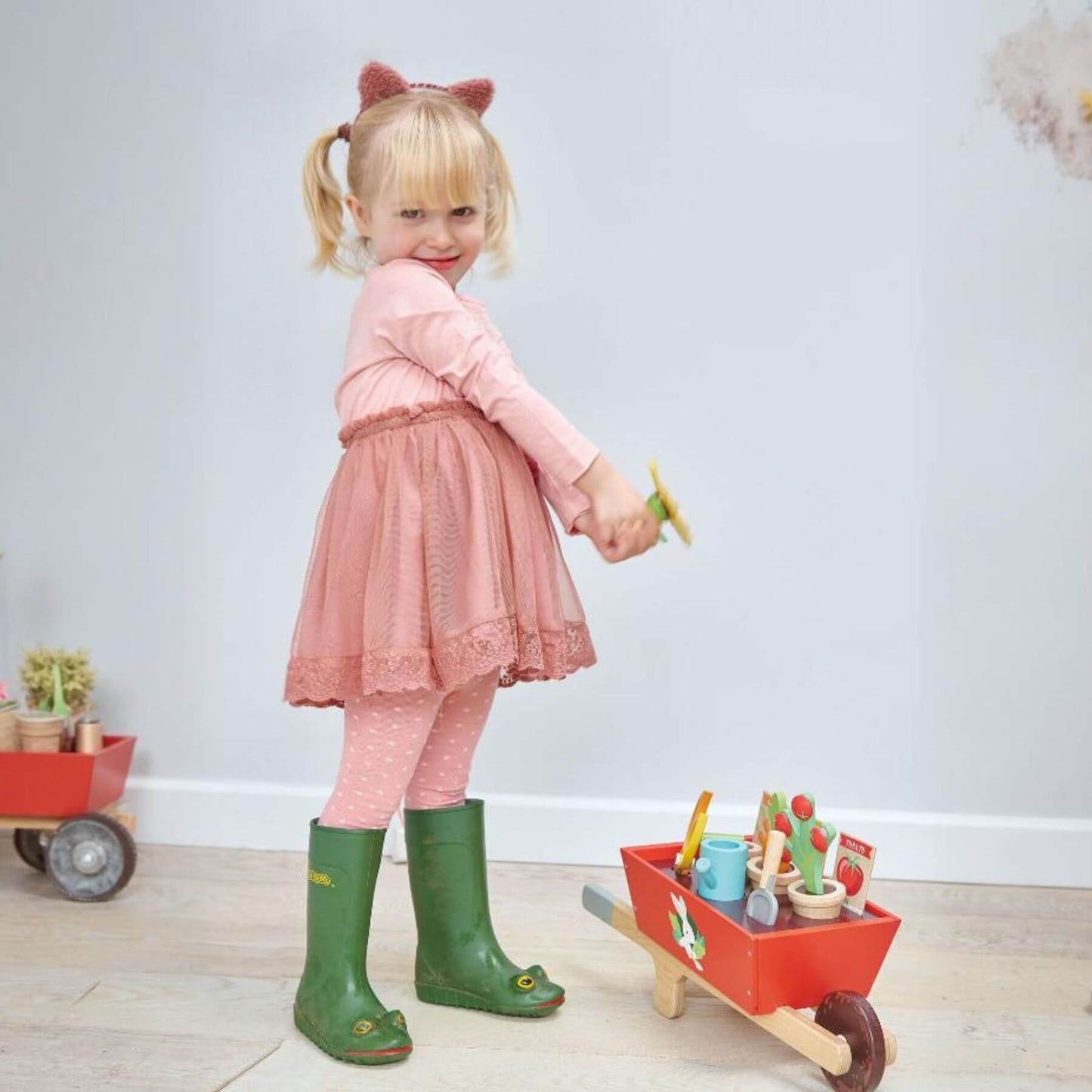 Child in pink dress and green boots playing with Tender Leaf Garden Wheelbarrow Set indoors.