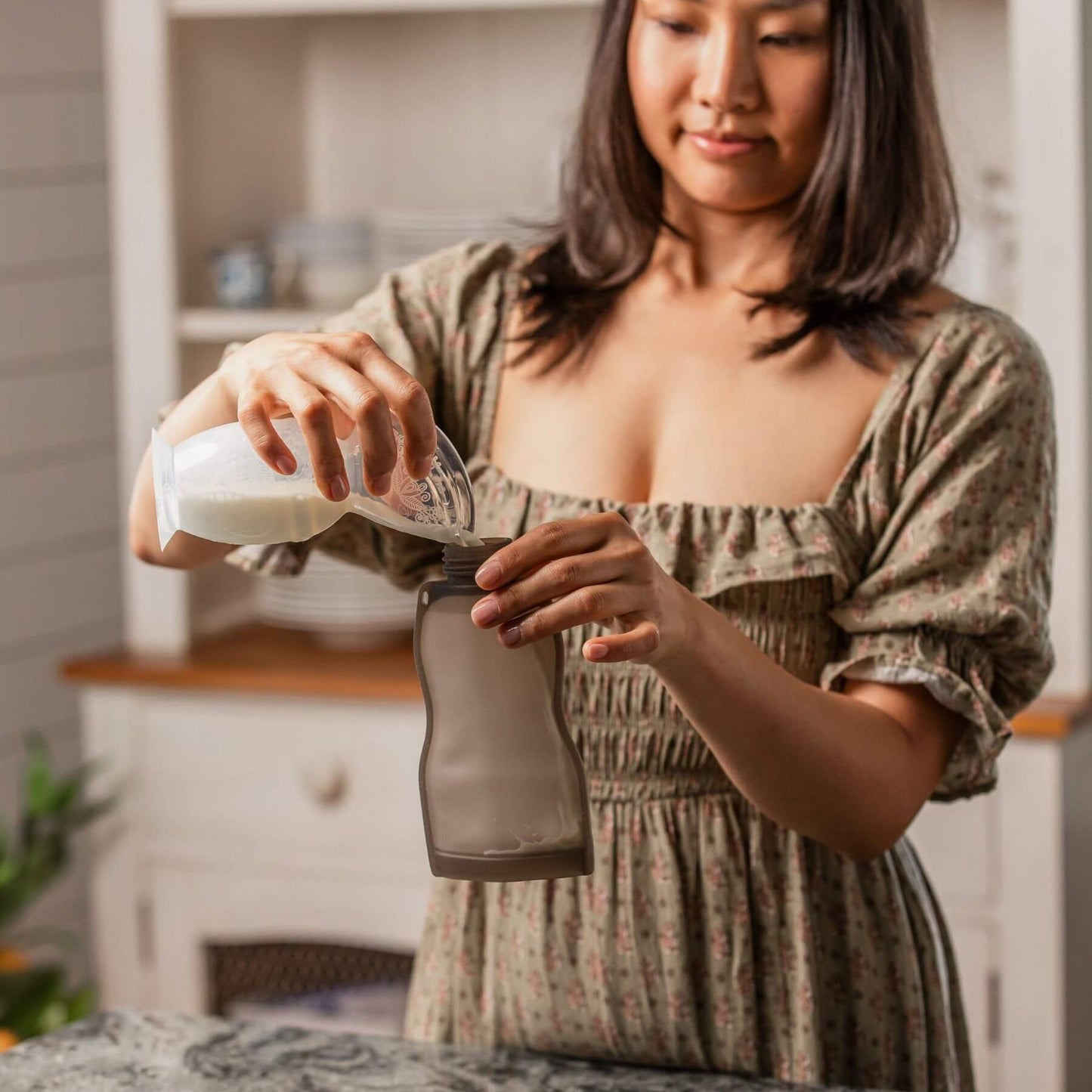 A woman in a floral dress pours liquid from a white bottle into a dark gray reusable bottle in a bright kitchen setting.