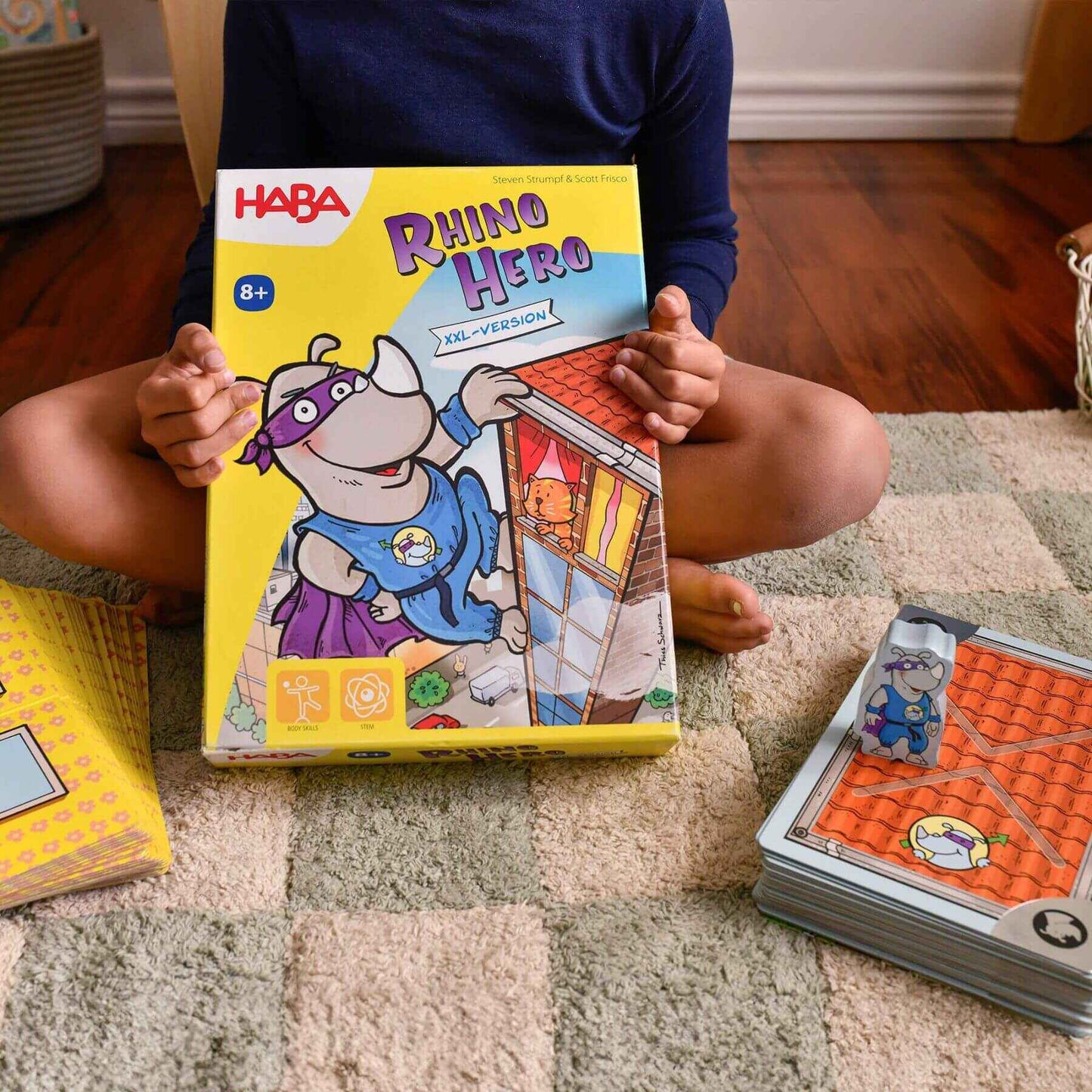 Child holding a HABA Rhino Hero game box on a carpeted floor with game cards nearby.