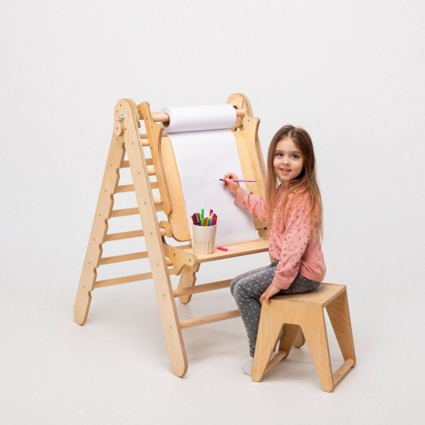 Child sitting on a wooden stool using a children's art easel with paper and coloring supplies.