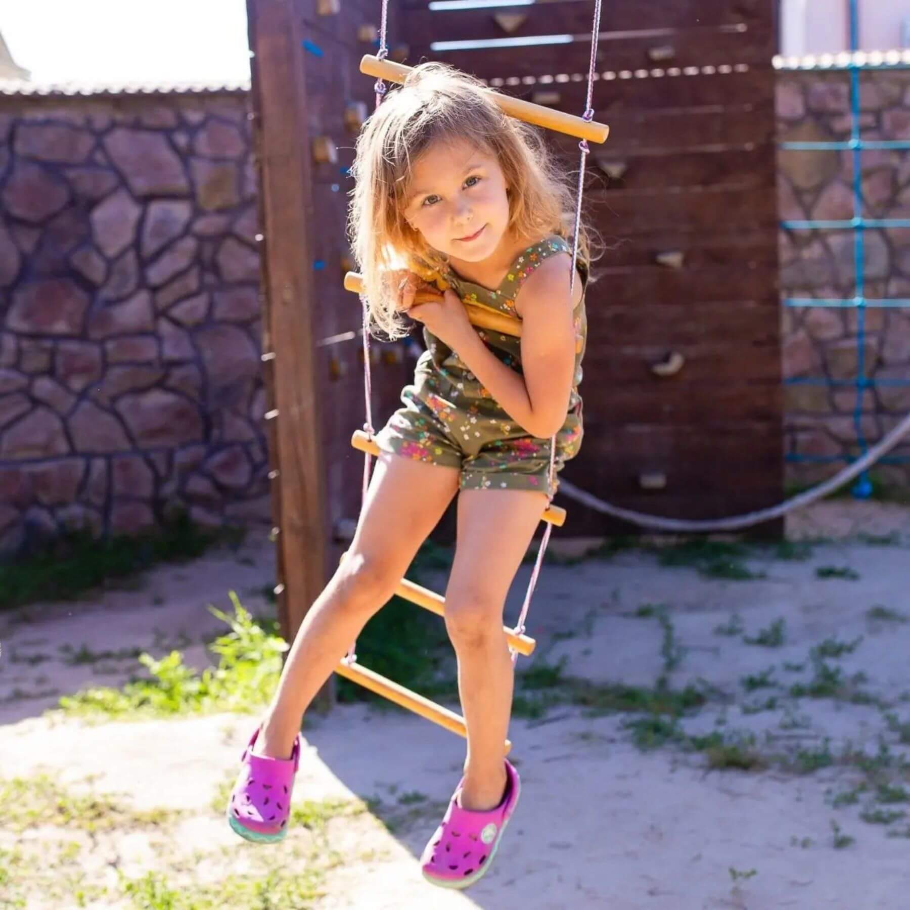 Young girl on Goodevas Climbing Rope Ladder with a stone wall and wooden structure.
