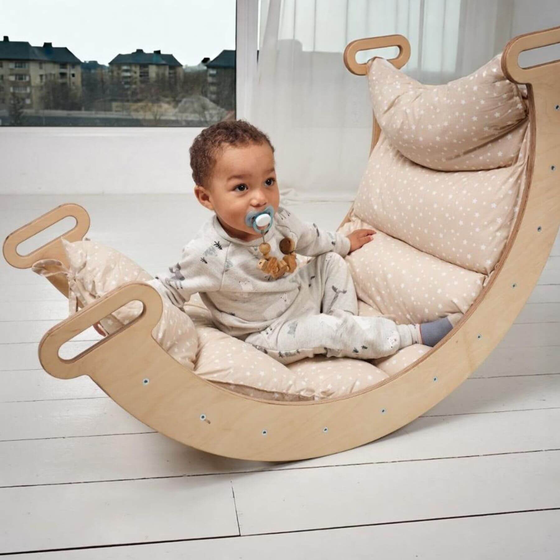 A baby sits in a curved wooden rocking chair with thick beige and white polka-dot cushions, set in a bright room with white floorboards and a large window.