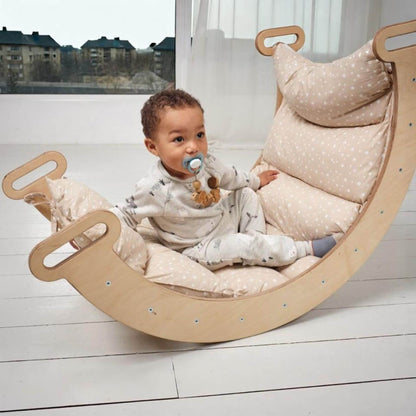 A baby sits in a curved wooden rocking chair with thick beige and white polka-dot cushions, set in a bright room with white floorboards and a large window.