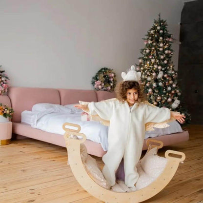 Child in a white onesie sitting on a wooden rocker in a room with a Christmas tree and pink bed.