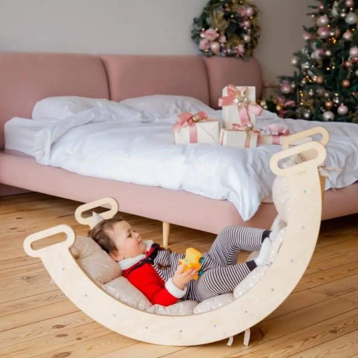 A toddler sits in a crescent-shaped wooden rocking swing with cushioned interior and loop handles, placed on a wooden floor in front of a pink bed with white bedding and Christmas decorations.