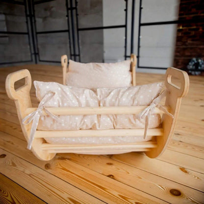 A wooden baby rocking cradle with a light beige dotted fabric cushion and matching pillow, set on a wooden floor in an indoor studio environment.