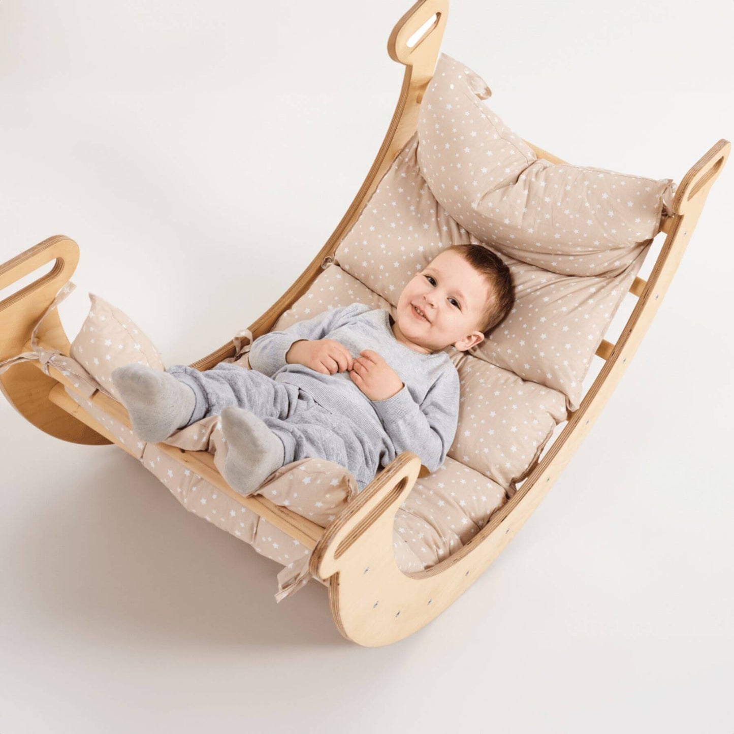 A baby lies on a padded wooden rocking chair with beige and white polka-dot cushions, photographed against a clean white background.