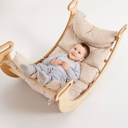 A baby lies on a padded wooden rocking chair with beige and white polka-dot cushions, photographed against a clean white background.