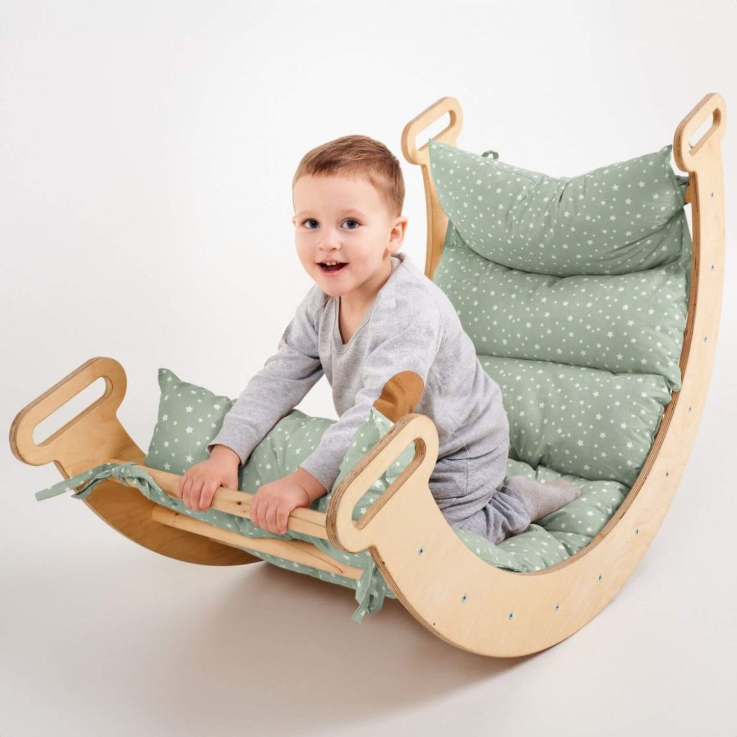 A smiling toddler sits on a wooden rocking chair with soft sage green cushions dotted with small white stars, photographed against a clean white studio background.
