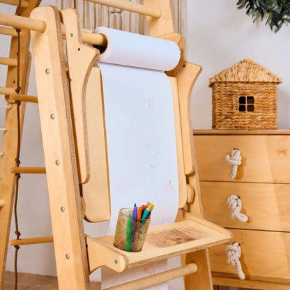 Wooden children's easel with paper roll, pencils, and a small table against a white wall.