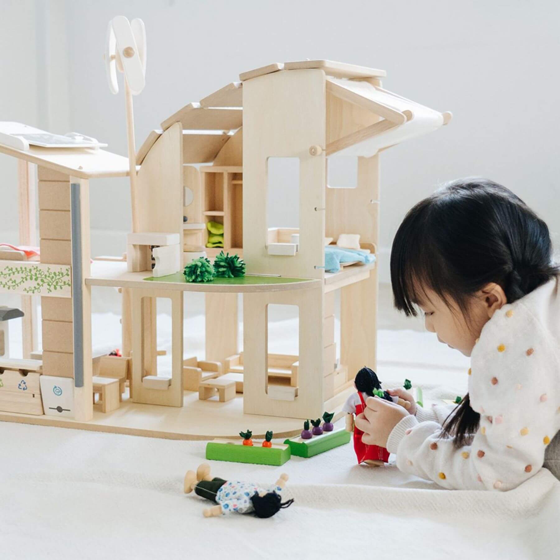 Child playing with PlanToys Green Dollhouse With Furniture and toys on a light-colored floor.