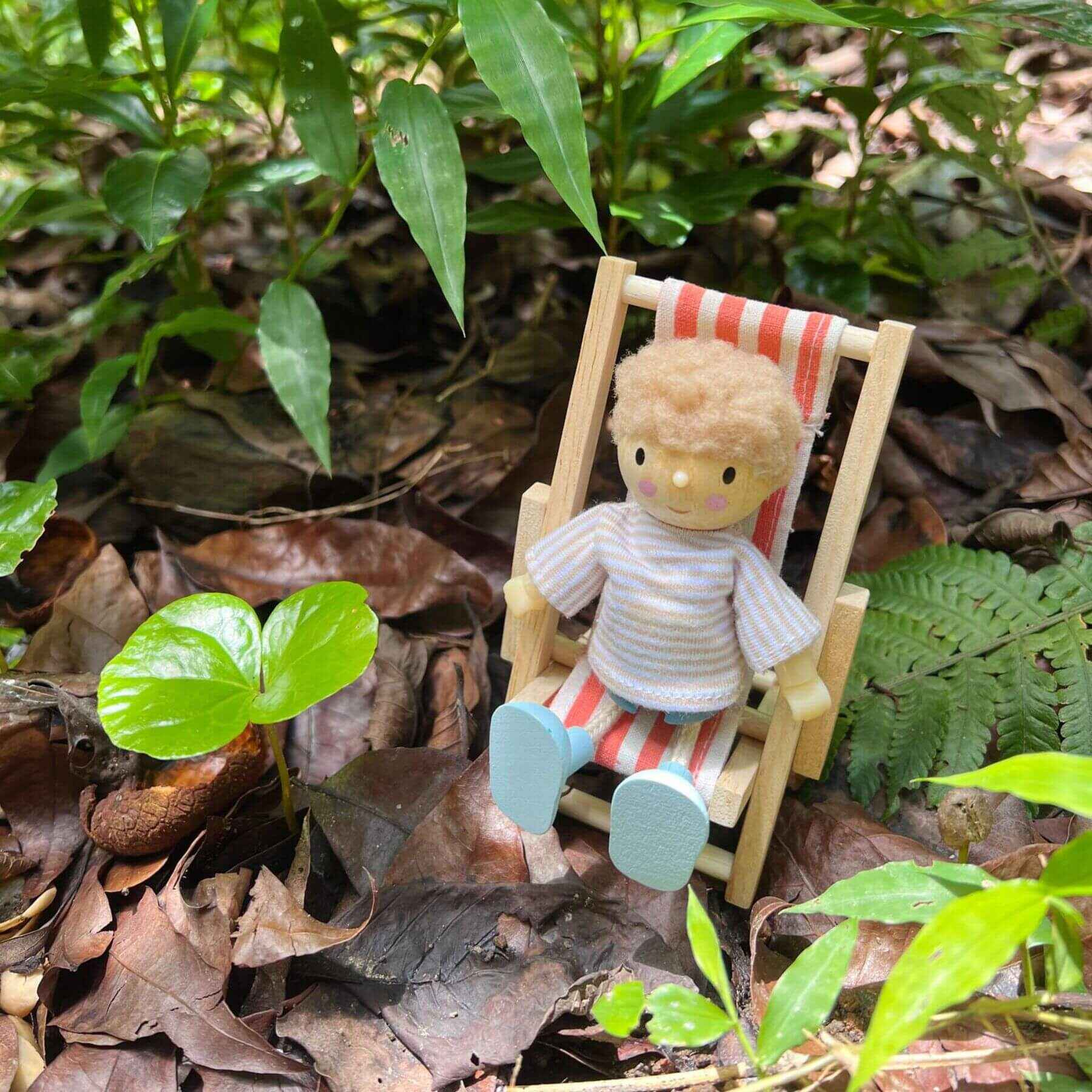 Small figurine of a person sitting in a striped chair among green leaves and brown leaves on the ground.