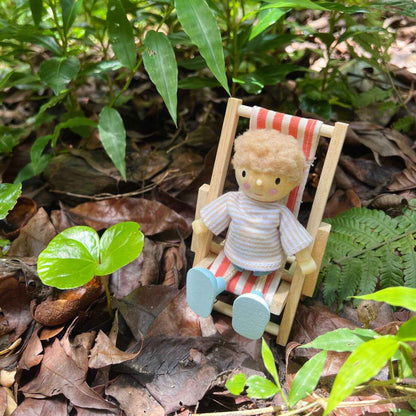 Small figurine of a person sitting in a striped chair among green leaves and brown leaves on the ground.