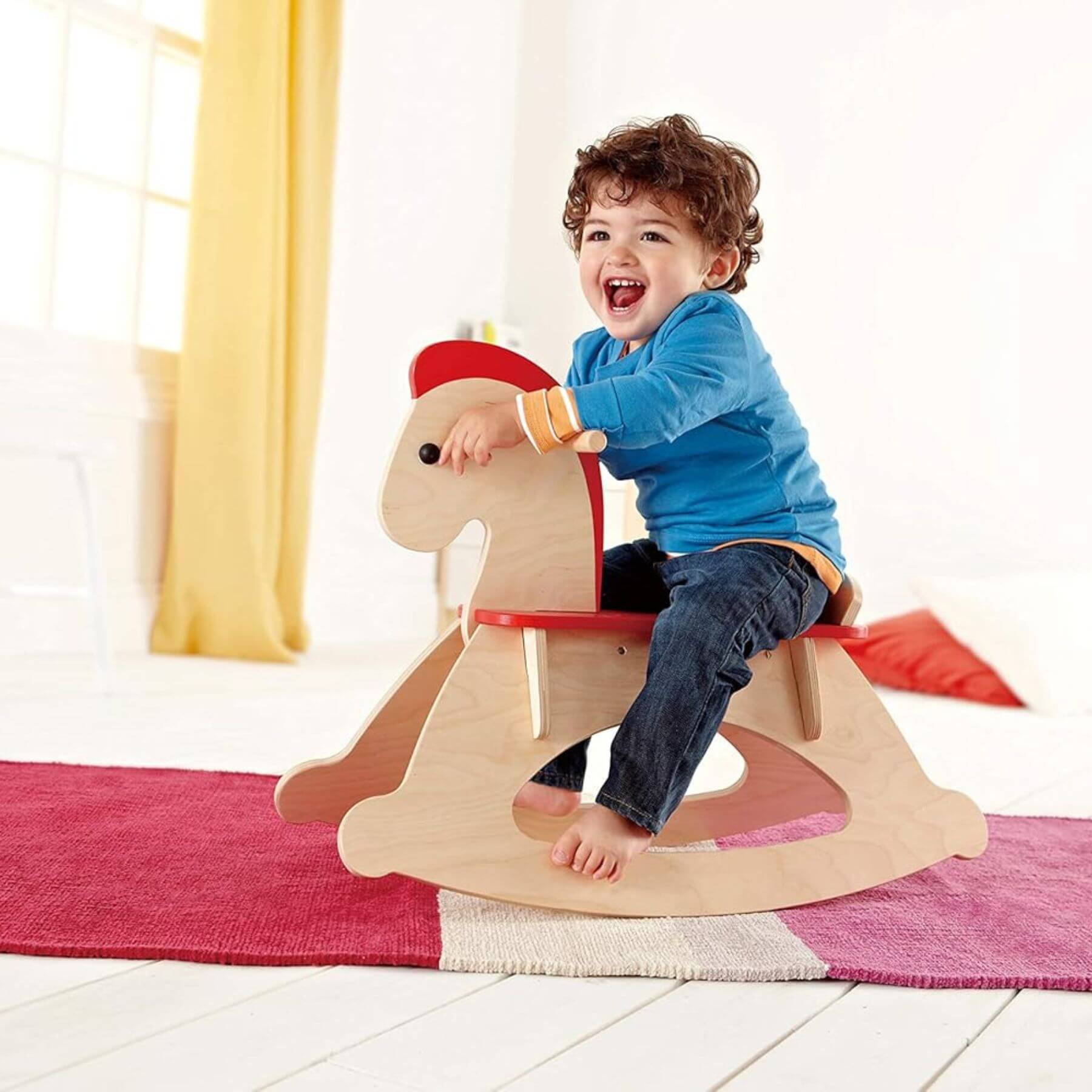 Child sitting on Hape Grow-with-me Rocking Horse in a bright room with yellow curtains.