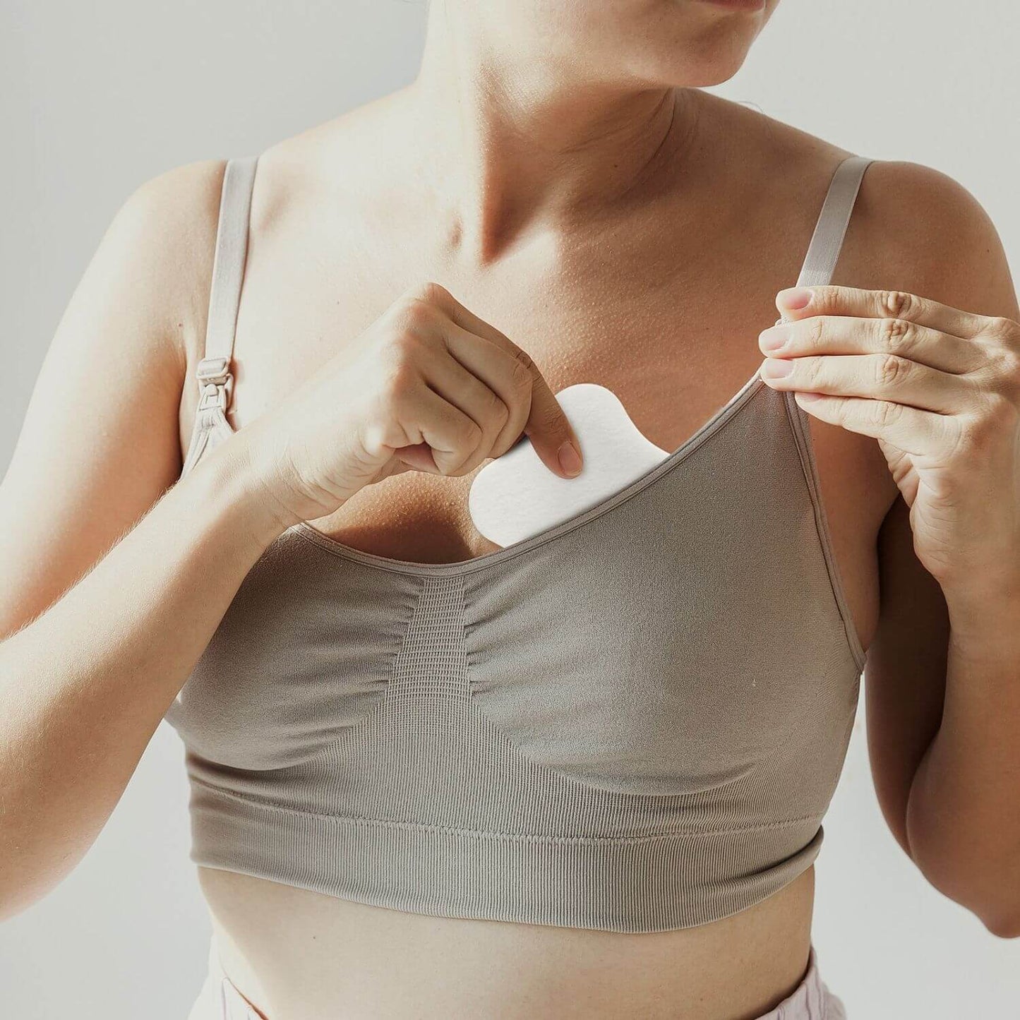 A close-up of a woman wearing a ribbed beige nursing bra and placing a white nursing pad into the front opening, with her hands adjusting the bra straps.