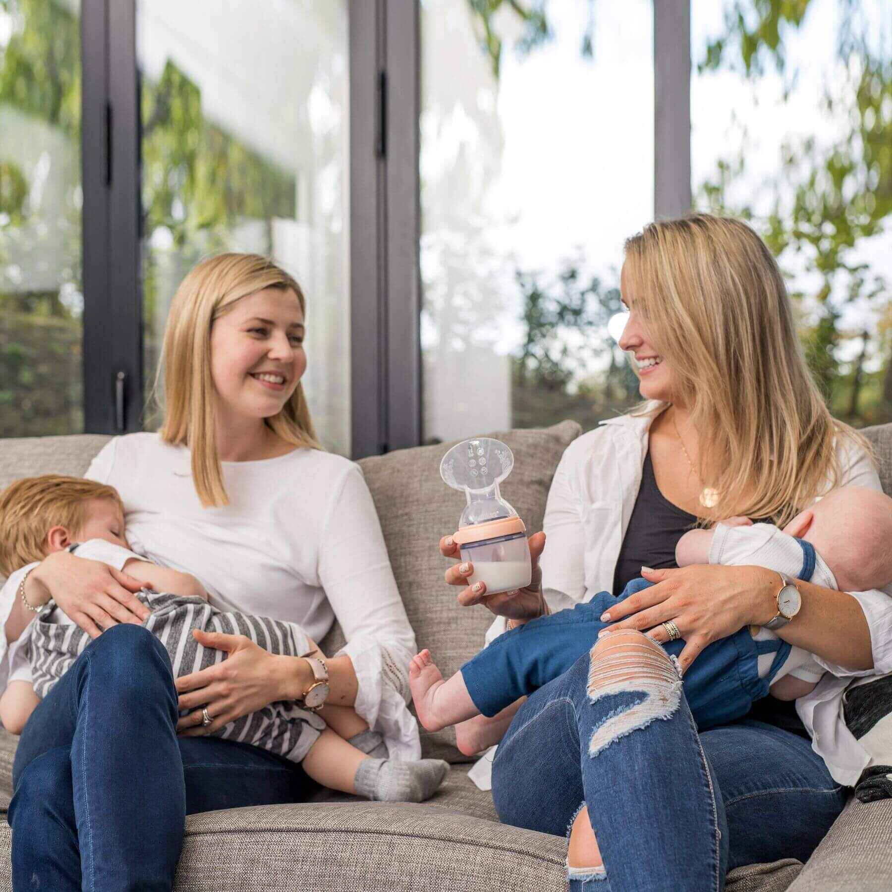 Two women sitting on a couch with babies, one holding a breast pump.