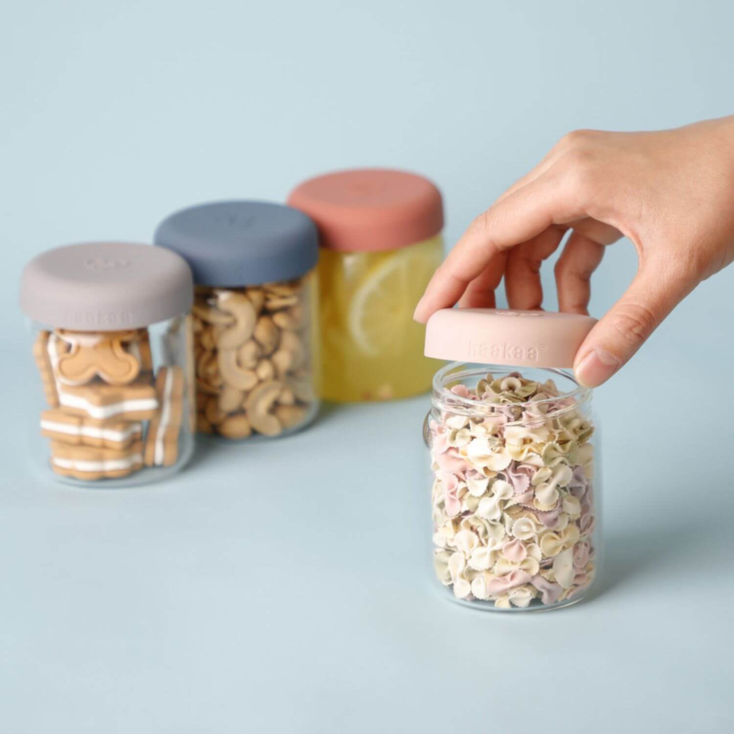 A hand is holding a pink lid on Haakaa Sealed Glass Storage Jar, with three more glass storage jars in the background containing snacks and drinks on a light blue surface.