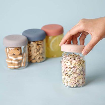A hand is holding a pink lid on Haakaa Sealed Glass Storage Jar, with three more glass storage jars in the background containing snacks and drinks on a light blue surface.