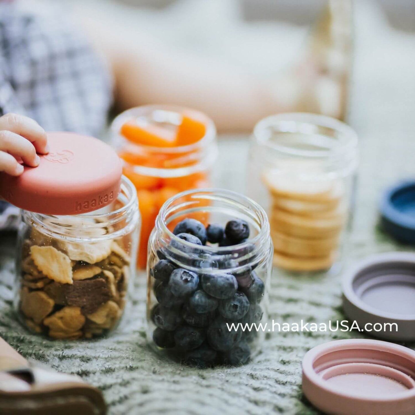 Haakaa Sealed Glass Storage Jar Set on a textured green blanket. A hand is holding the lid of a jar filled with dried fruit and crackers, with other jars containing blueberries, carrot sticks, and cookies in the background.
