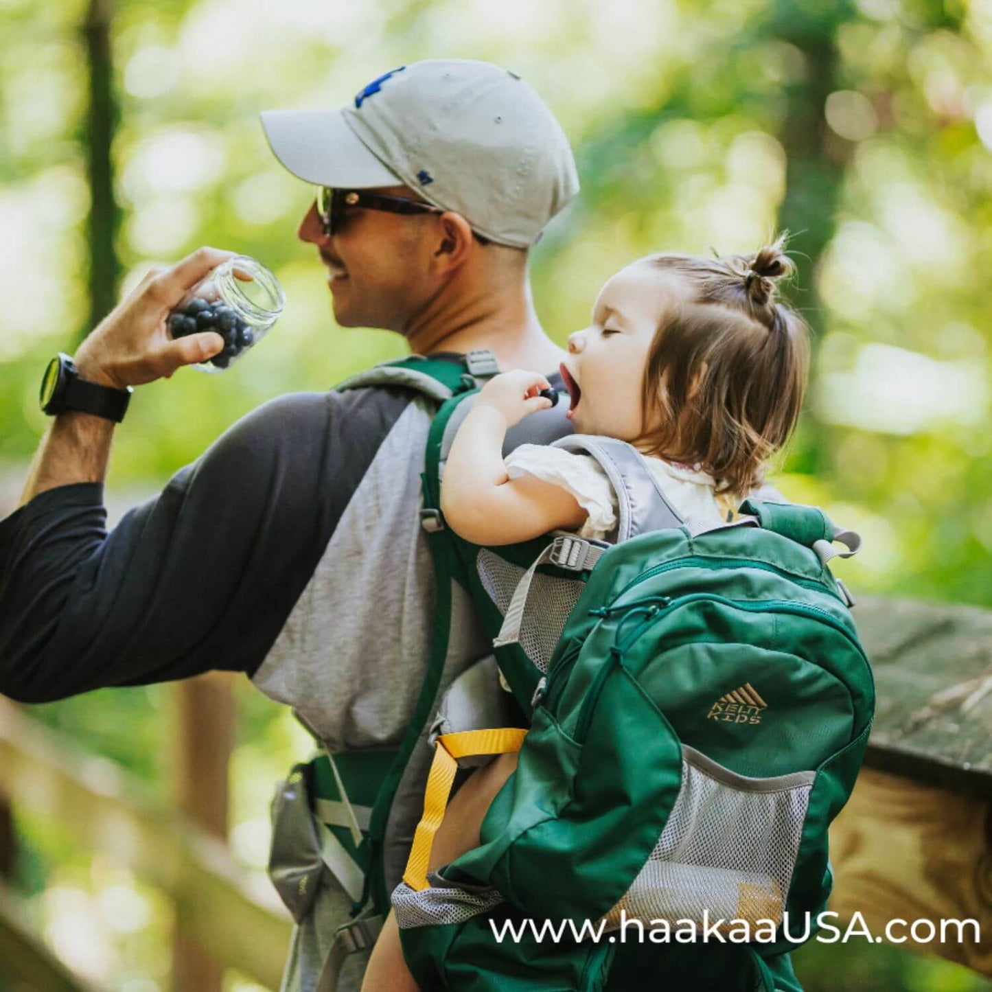 A man hiking outdoors with a child in a green Haakaa baby carrier backpack. The man is holding a Haakaa Sealed Glass Storage Jar, and the child is sitting in the carrier.