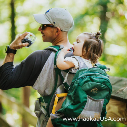 A man hiking outdoors with a child in a green Haakaa baby carrier backpack. The man is holding a Haakaa Sealed Glass Storage Jar, and the child is sitting in the carrier.