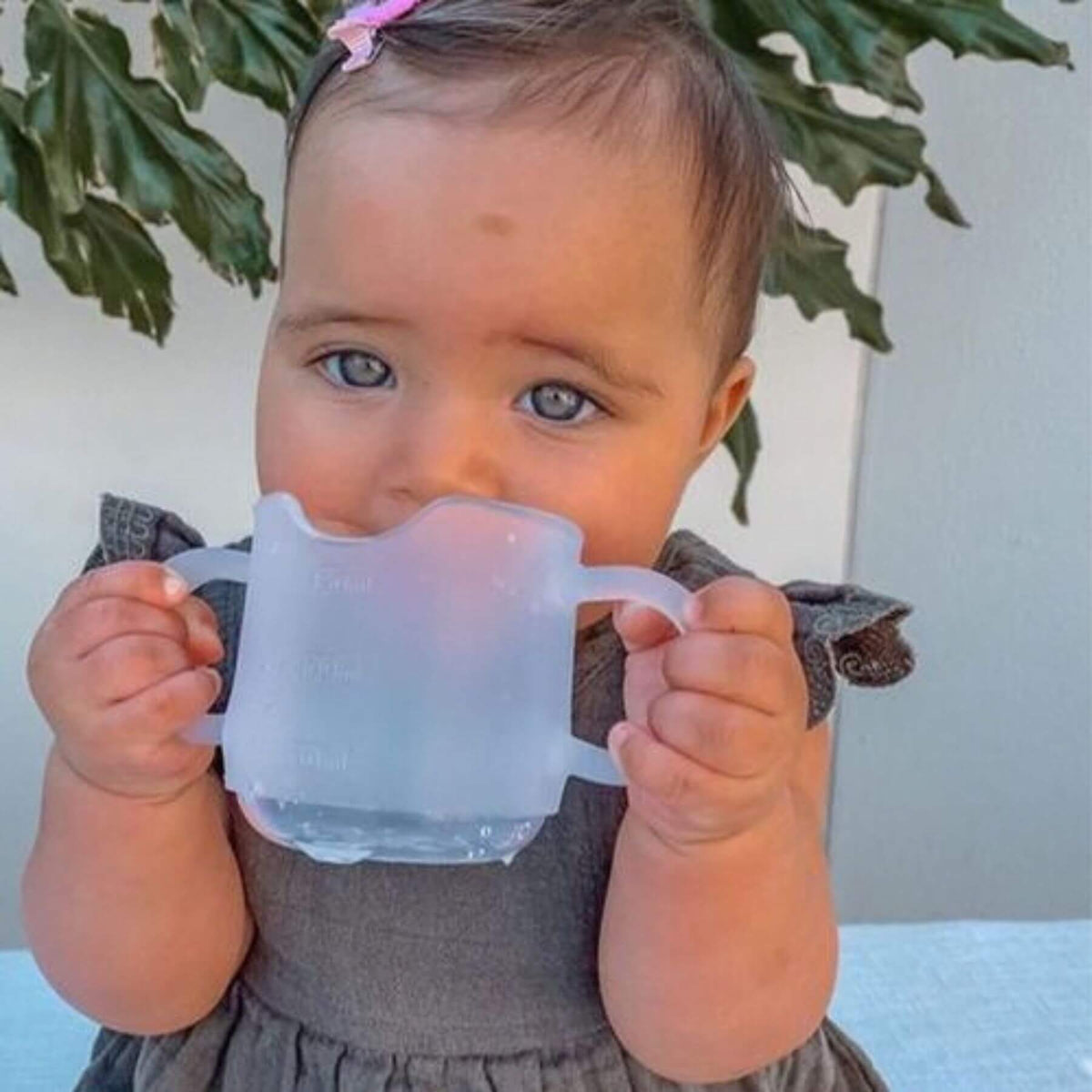 Baby holding Haakaa Silicone Baby Drinking Cup with a plant in the background