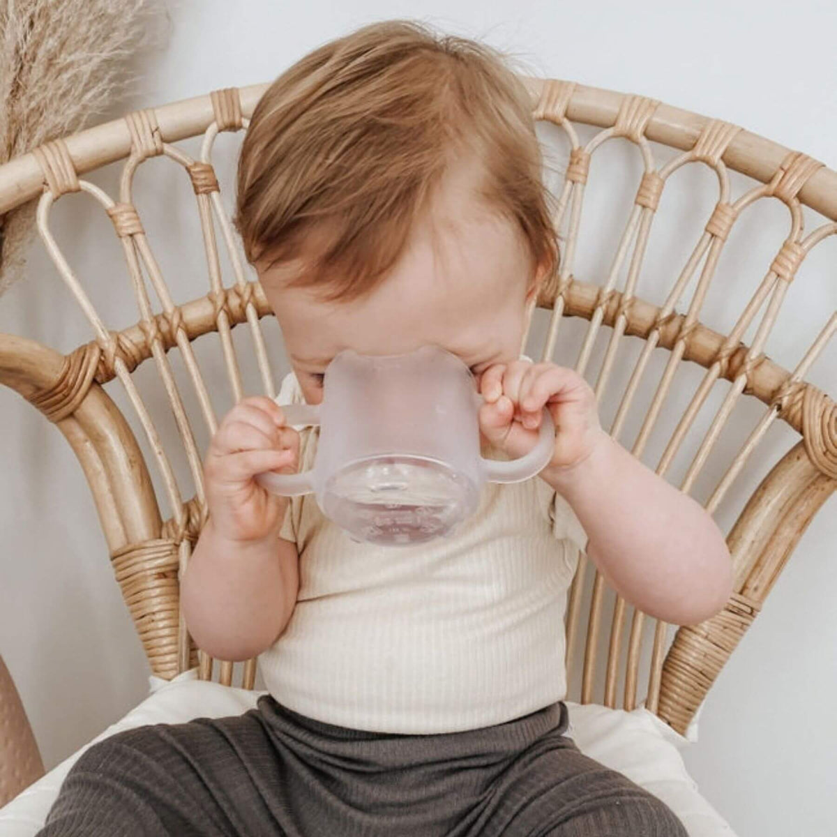 A baby sitting in a woven rattan chair drinking from Haakaa Silicone Baby Drinking Cup