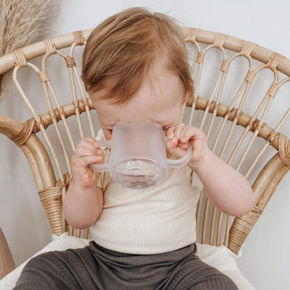 A baby sitting in a woven rattan chair drinking from Haakaa Silicone Baby Drinking Cup