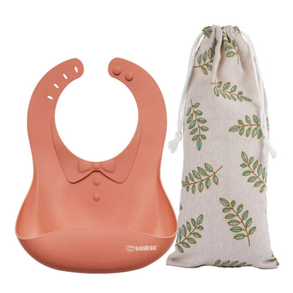 Haakaa Silicone Baby Bib - Rust with a bow tie and a drawstring bag with leaf pattern on a white background