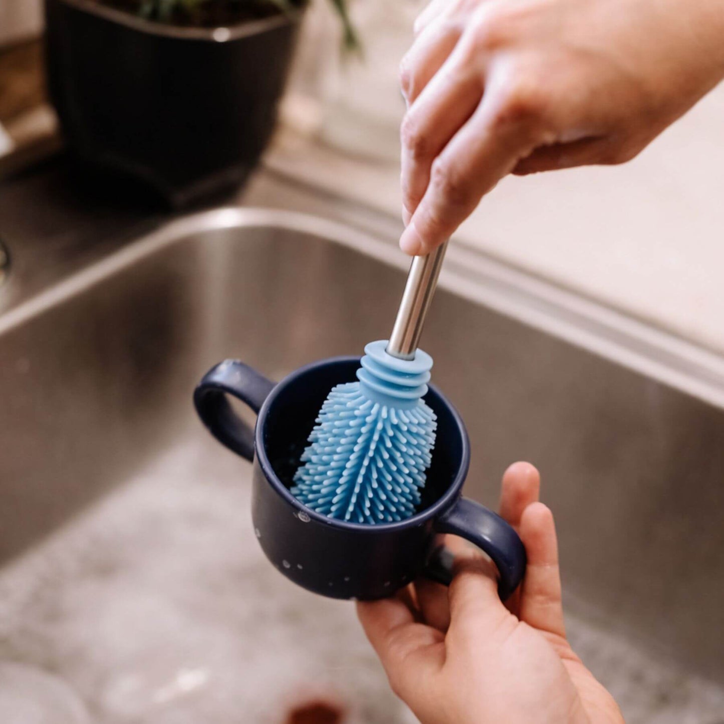 Person using a blue silicone scrubber in a black mug by a sink.