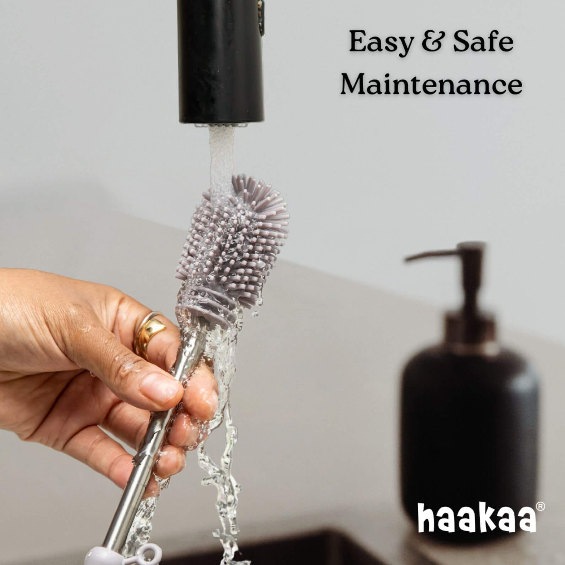 Hand holding a cleaning brush with water flowing, next to a black haakaa product.