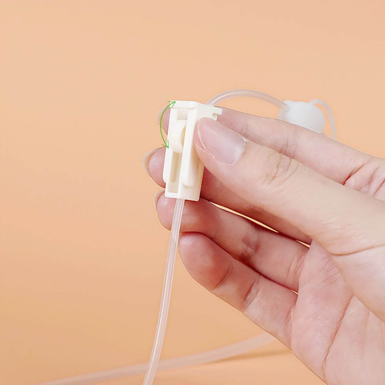 A hand holding a small white plastic tube connector with a clear tubing piece and a thin green wire, against a soft peach background.