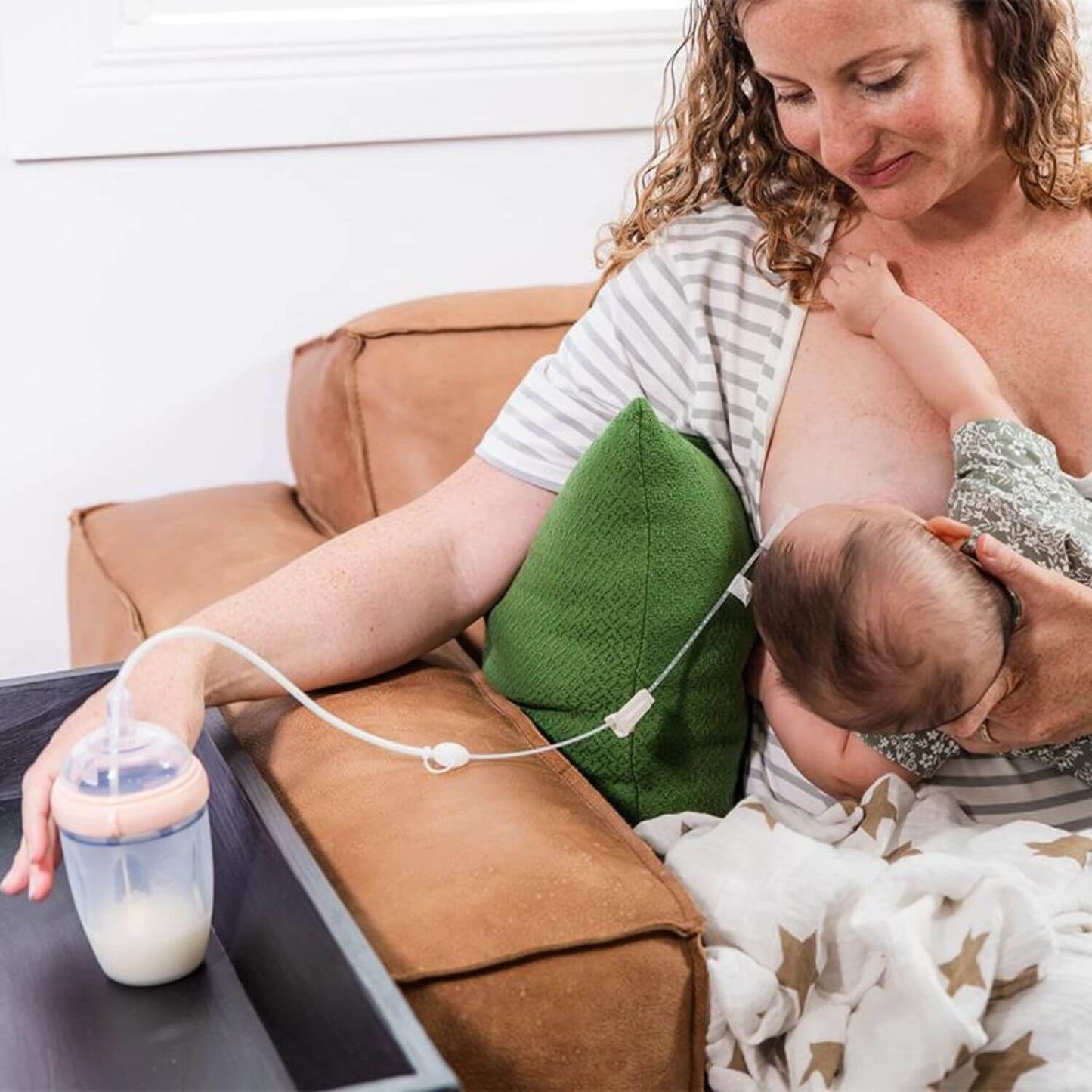 A woman breastfeeding a baby while using Haakaa Supplemental Feeding Combo resting on a black tray next to a brown leather couch.